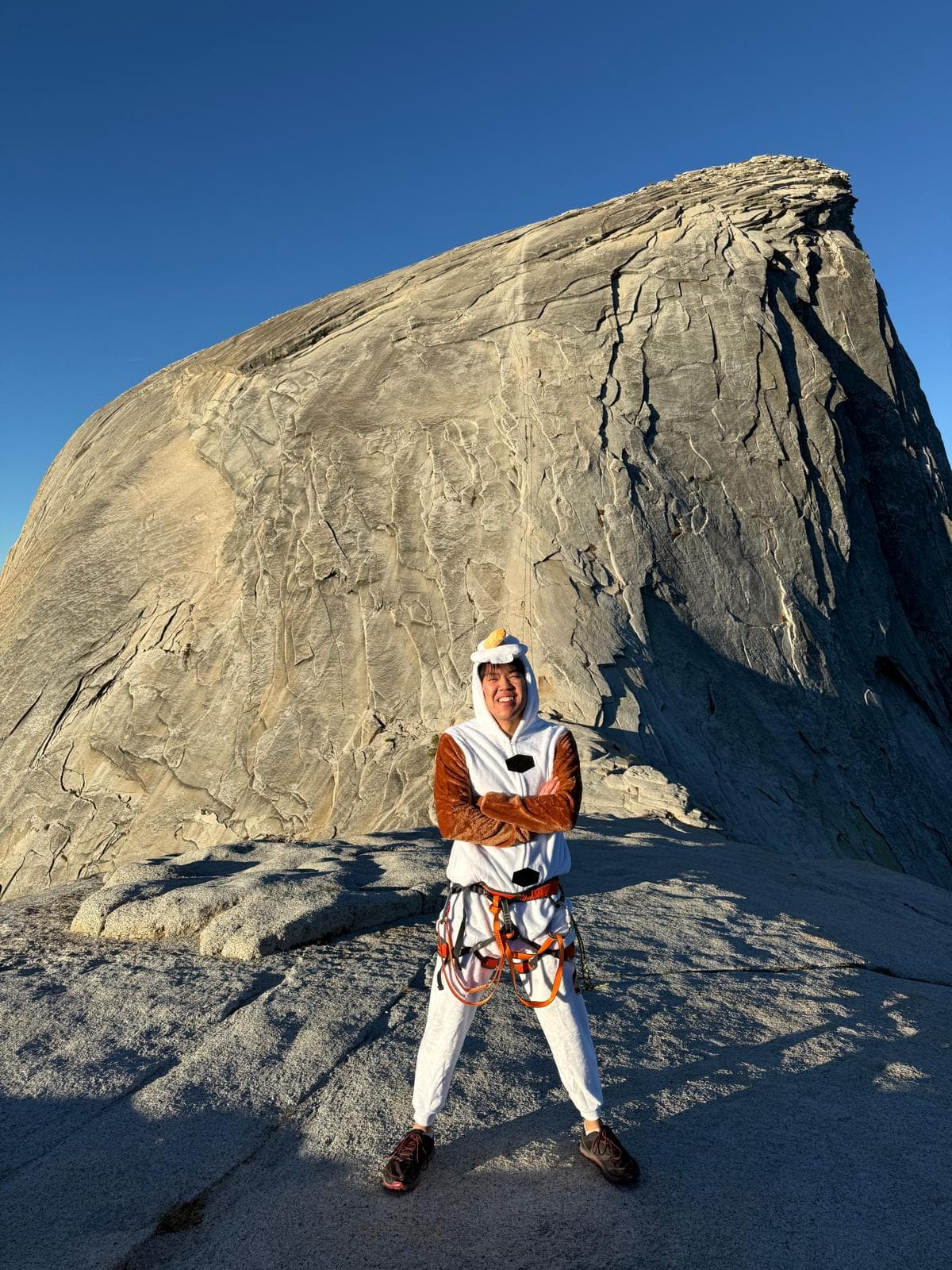 Jethro Chu on the granite summit of Half Dome at golden hour, in a climbing harness and a white costume, arms crossed and smiling.